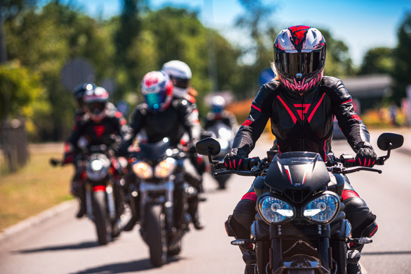 grupa motocyklistów jedzie w pięknej pogodzie, przewodzi im kobieta w czarno-różowym kombinezonie Rebelhorn