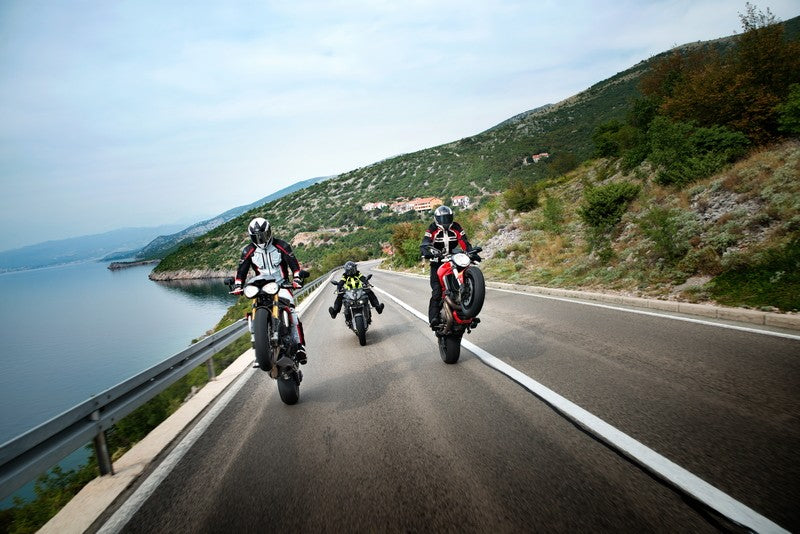 Trójka motocyklistów jedzie drogą wzdłuż morza, dwóje z nich jedzie na tylnym kole