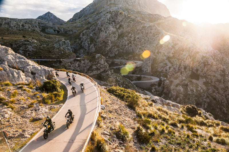Trasa motocyklowa górach, na drodze kilku motocyklistów w kaskach, w tle zachodzące słońce