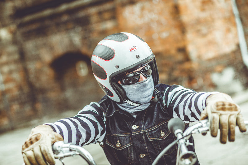 Motocyklista siedzący na motocyklu, w kasku Bell, ciemnych okularach i chuście motocyklowej, pasiasta motocyklowa koszulka i rękawice oparte na kierownicy motocykla