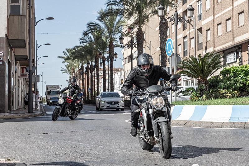 Motocyklista w czarnym kasku i kurtce motocyklowej skórzanej jadący ulicą miasta na motocyklu, w tle budynki, palmy i inny motocuuklista