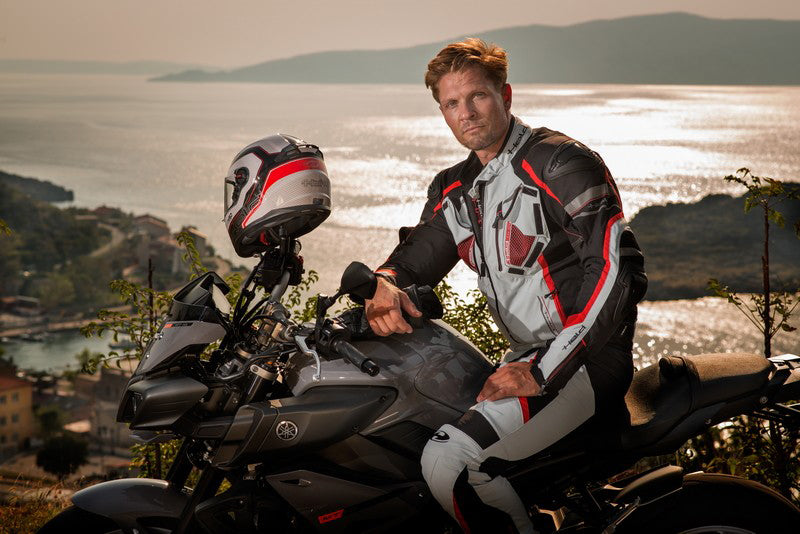 Mężczyzna pozujący na motocyklu w kombinezonie motocyklowym marki Held i z kaskiem motocyklowym zawieszonym na kierownicy motocykla, w tle morze 