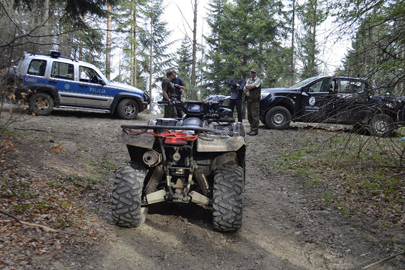 Quad stojący na leśnej drodze, przed nim cztery rozmawiające osoby, policyjne auto oraz samochód straży leśnej