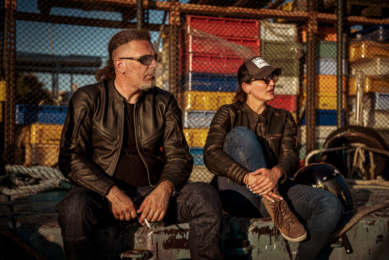 Mężyczna i kobieta w odzieży motocyklowej i okularach przeciwsłonecznych, siedzący na murku i patrzący w jednym kierunku