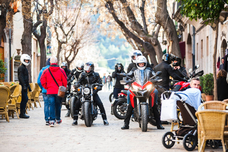Grupa motocyklistów w kaskach stojących na zatłoczonej uliczce, obok przechodnie i kawiarnie