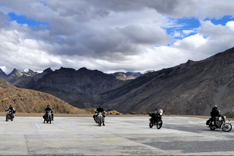 Motocykliści w turystyce motocyklowej, siedzący na motocyklach, w odległości od siebie, ubrani w kaski motocyklowe, w tle góry i niebo