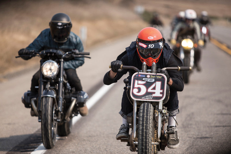 grupa motocyklistów na drodze w kaskach marki Bell. Jeden z motocyklistów ma tabliczkę na przodzie motocykla z numerek 54