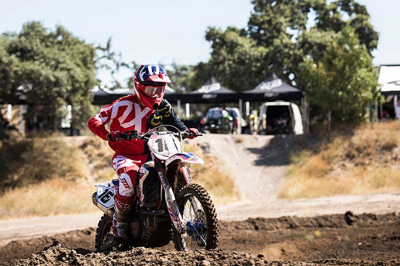 Motocyklista offroadowy w kombinezonie i kasku na motocyklu wjeżdża po hałdach piachu, w tle drzewa i niebo