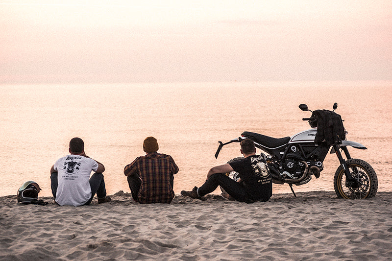 Trzech mężczyzn w ubraniach motocyklowych, obok motocykl i zachodzące słońce nad wodą