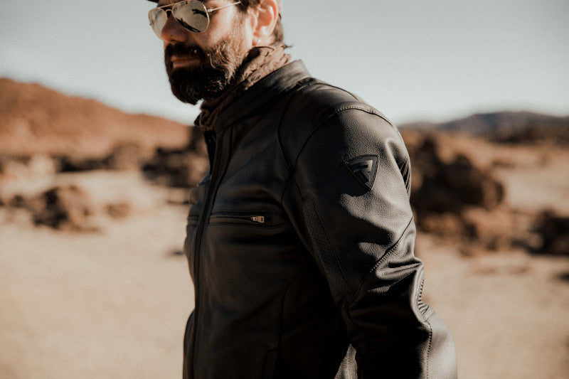 Mężczyzna z brodą pozujący w motocyklowej kurtce i okularach przeciwsłonecznych, w tle pustynia