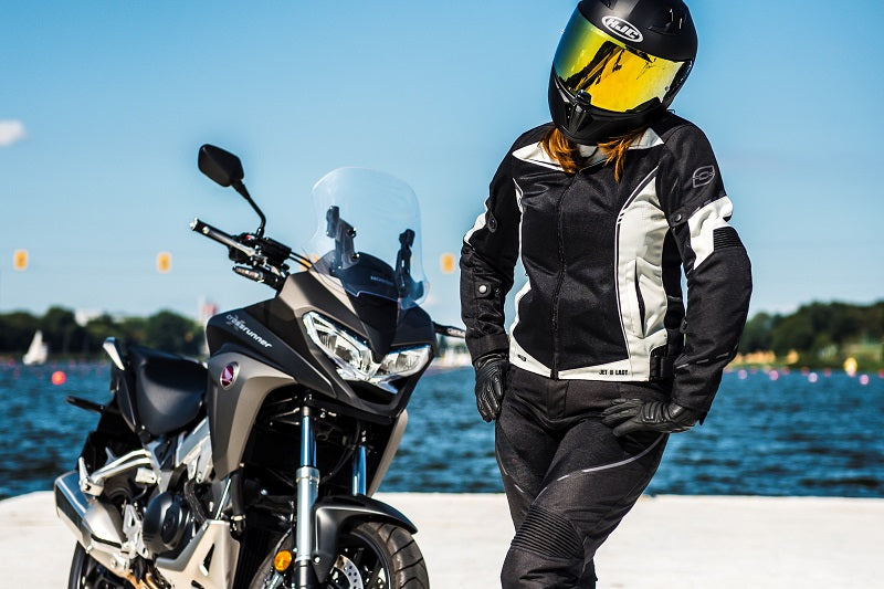 Motocyklista w damskiej kurtce motocyklowej rebelhorn i kasku, pozuje do zdjęcia obok motocykla, w tle woda