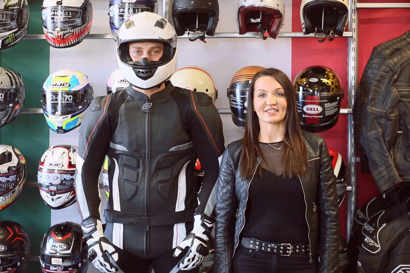 Prezentacja kasku motocyklowego na modelu ubranych w kask i strój motocyklowy, obok kobieta prezentująca strój, w tle ściana z kaskami