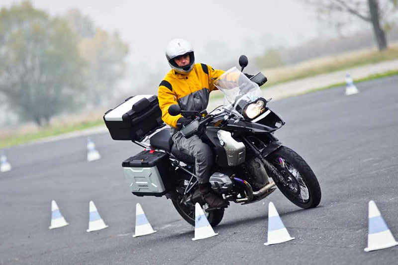 Motocyklista w żółtej kurtce i kasku na motocyklu z kuframi na bagażniku, przejeżdżający po placyku obok słupków.