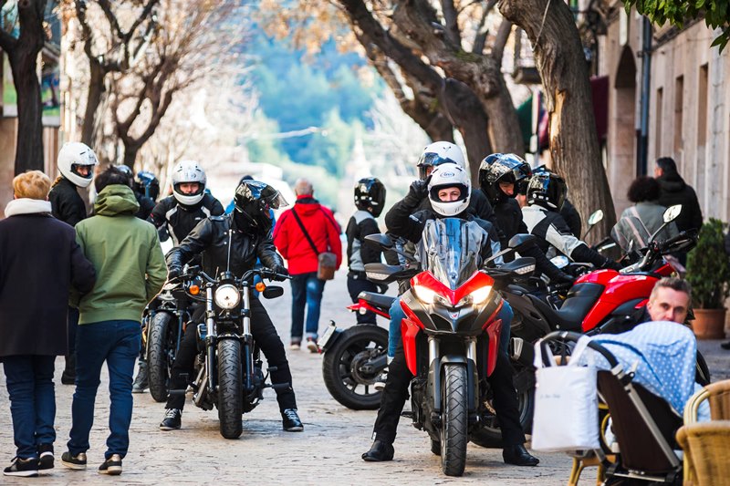 Grupa motocyklistów w kaskach stojących na zatłoczonej uliczce, obok przechodnie i kawiarnie