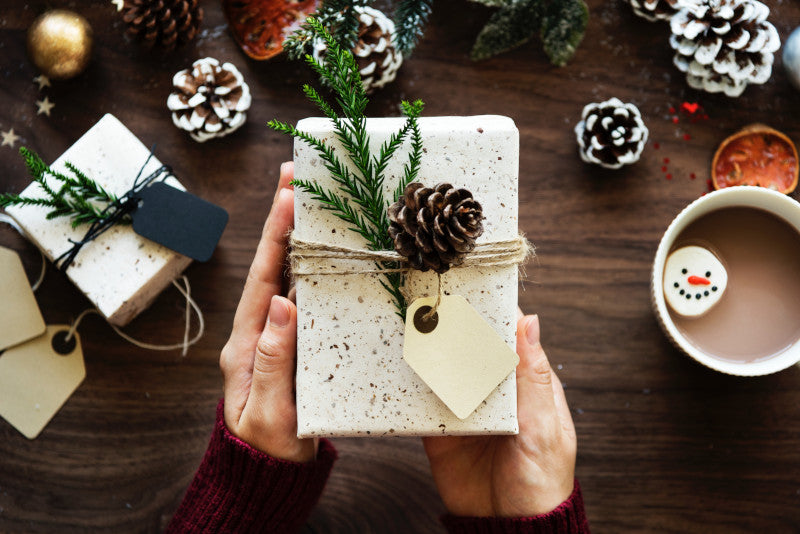 Świąteczny ozdobnie zapakowany prezent trzymany w dłoniach, gorące kakao i szyszki
