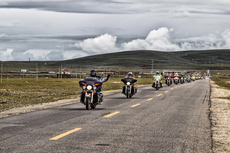 grupa motocyklistów jedzie drogą wśród wzgórz i pozdrawia fotografa ręką