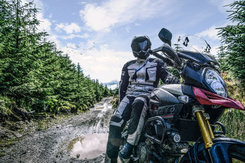 Motocyklista pozujący przy motocyklu, obrany w kombinezon motocyklowy marki RST i kask motocyklowy, w tle strumyk, błoto i las.