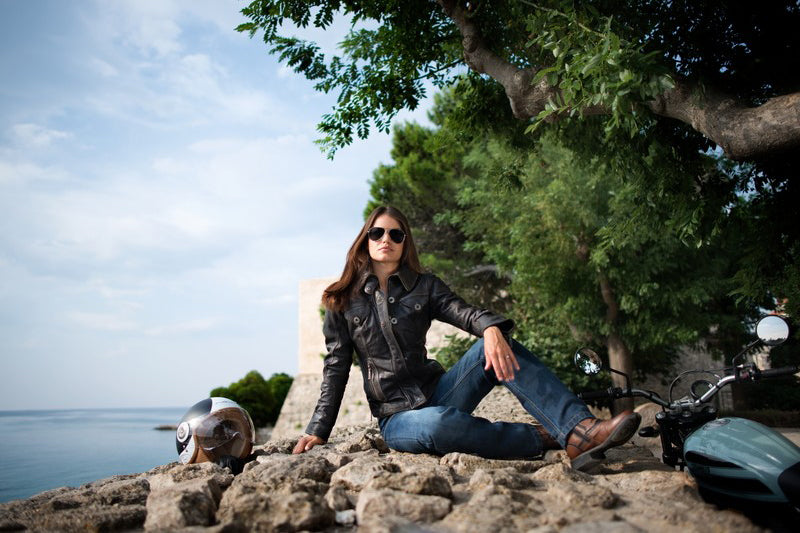 Kobieta siedząca na skale obok drzew, w tle woda, ubrana w jeansy, skórzaną kurtkę motocyklową i ciemne okulary, obok niej kask i motocykl 